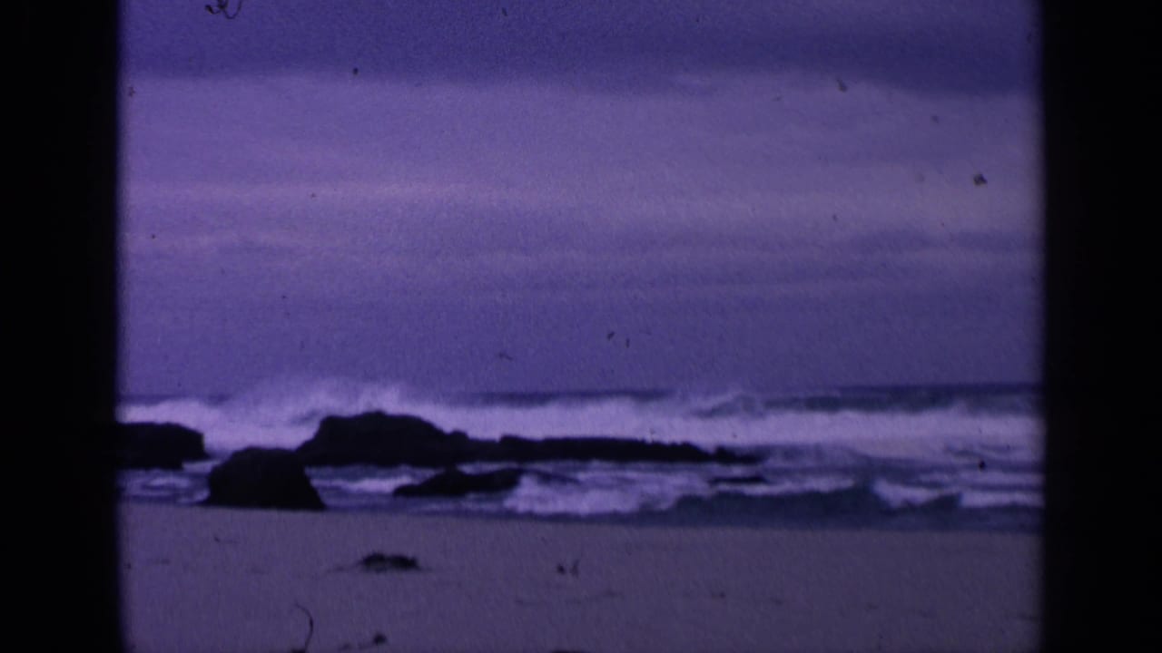 1963 San Francisco 8mm frame showing a purple-blue rocky beach with low waves, sand, and a distant ocean horizon.