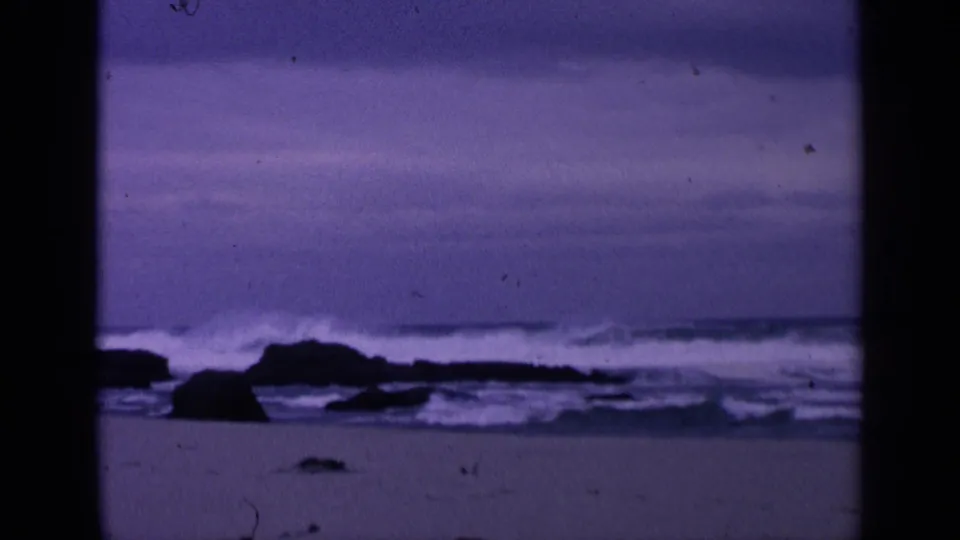 1963 San Francisco 8mm frame showing a purple-blue rocky beach with low waves, sand, and a distant ocean horizon.