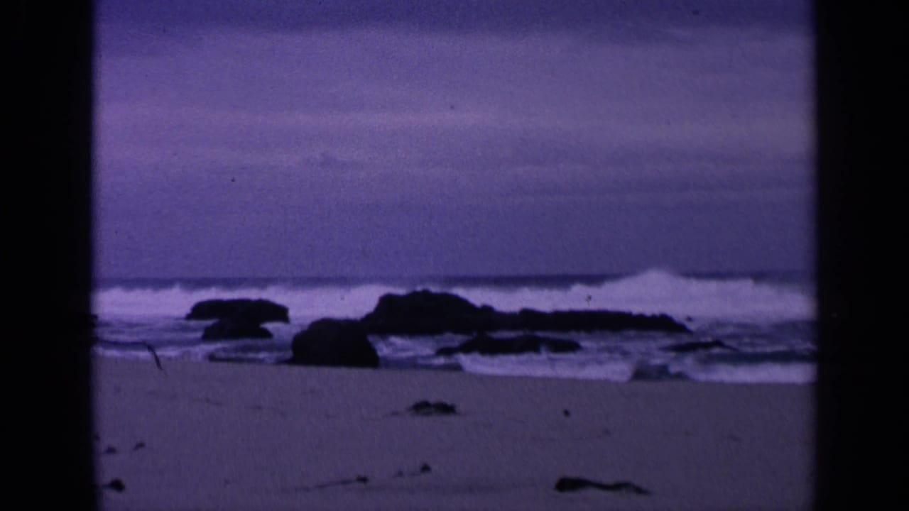 1963 San Francisco coastal home-movie frame with surf breaking around dark rocks beneath a purple-blue sky.