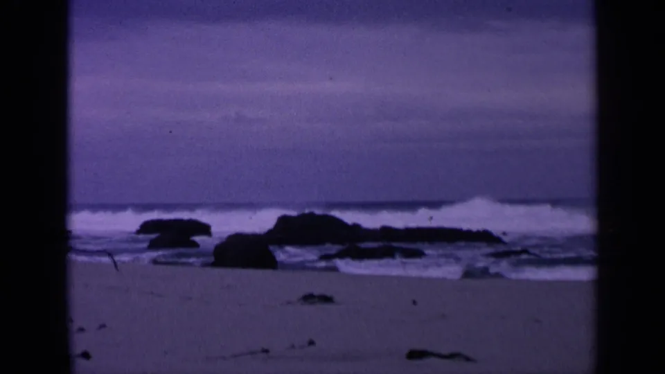 1963 San Francisco coastal home-movie frame with surf breaking around dark rocks beneath a purple-blue sky.