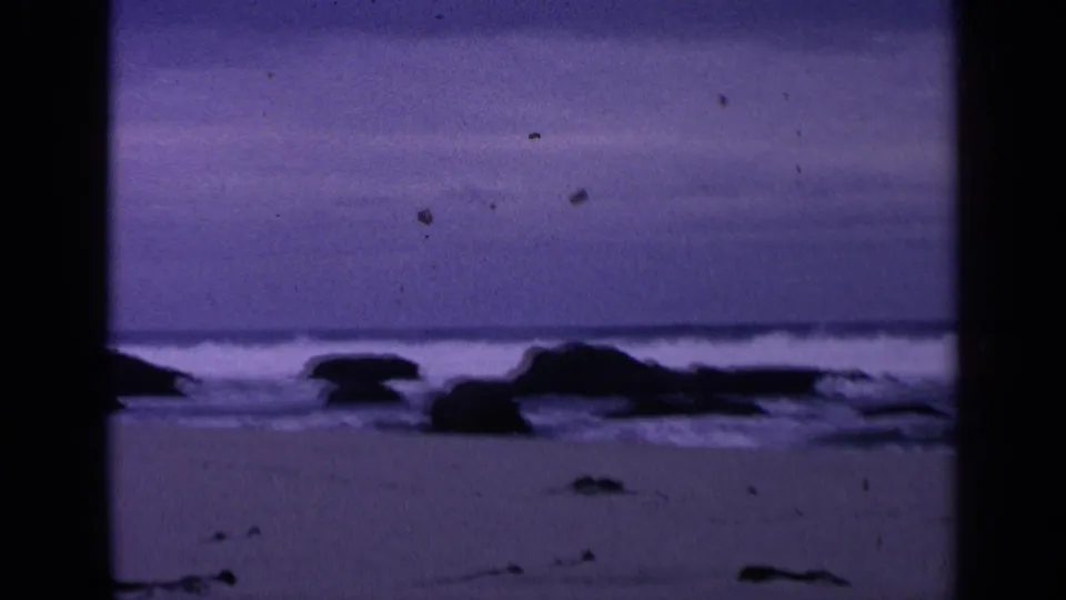 1963 San Francisco 8mm still of a sandy rocky shoreline with low waves and a strong purple film tint.