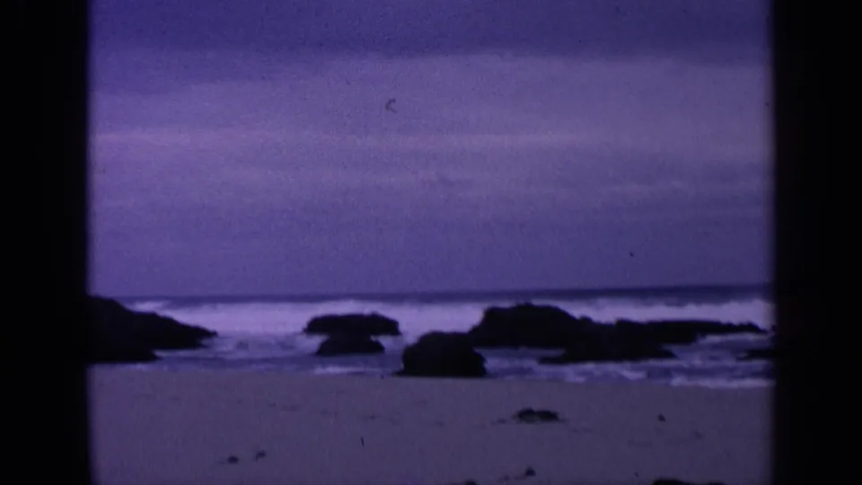 1963 San Francisco home-movie frame showing a purple-cast beach, scattered rocks, and surf near the horizon.