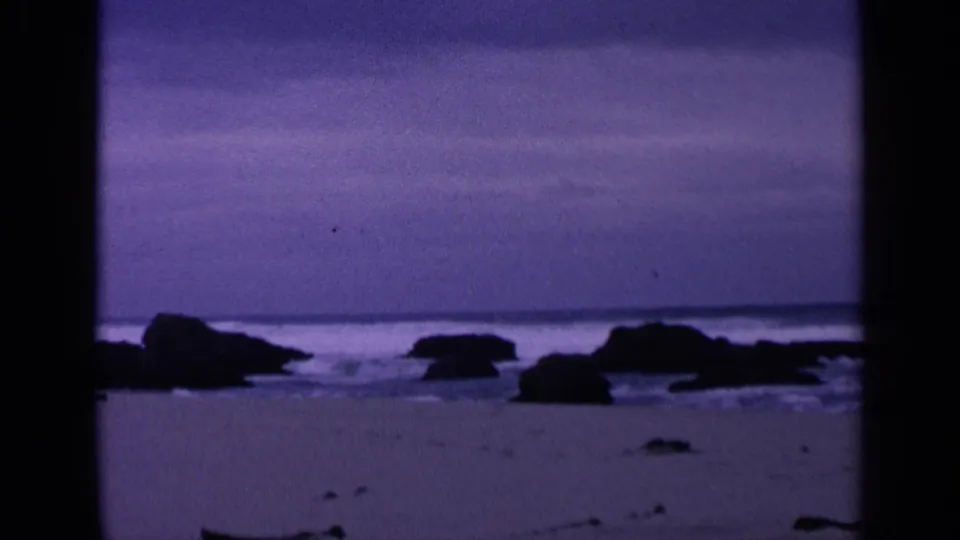 1963 8mm home-movie view of a San Francisco shoreline with low surf, scattered dark rocks, wet sand, and a purple-blue sky.