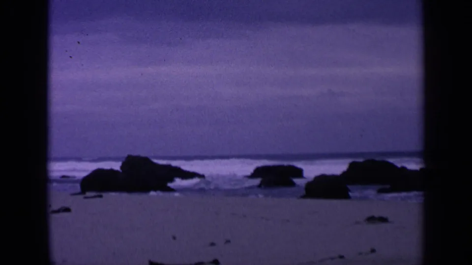 San Francisco coastal home-movie frame from 1963 showing small waves, dark shoreline rocks, and a muted purple sky over wet sand.