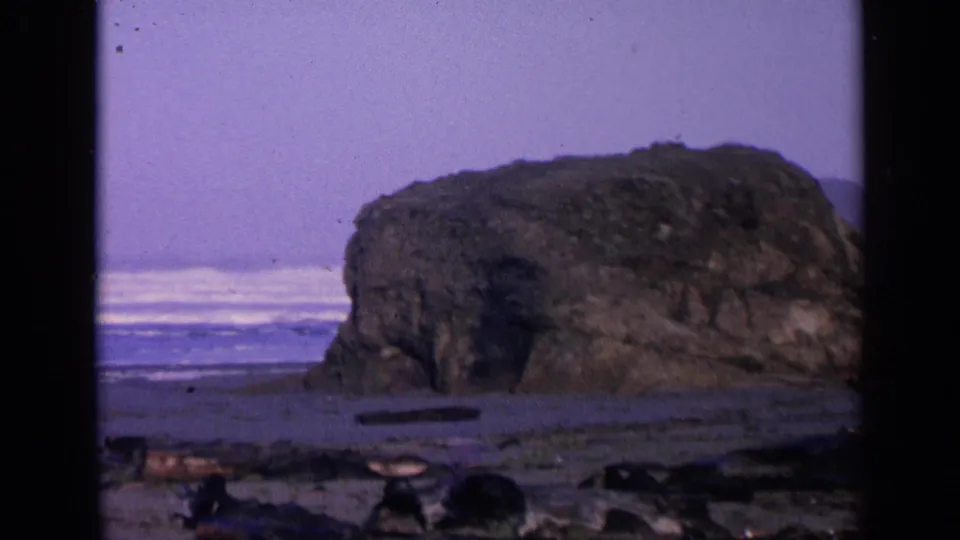1963 San Francisco beach footage frame with a large sea rock in the foreground area, surf beyond it, and scattered beach debris or stones.