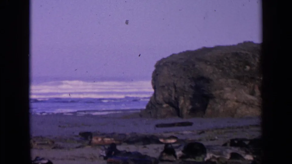 San Francisco 1963 8mm frame showing a large shoreline boulder, foamy waves in the background, wet sand, and purple-blue film color.