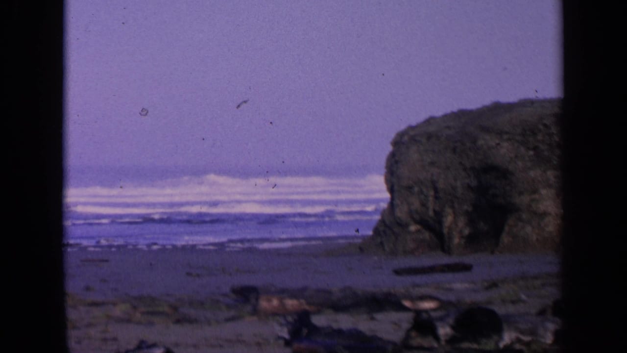 1963 San Francisco-area 8mm frame showing Pacific surf, sandy shoreline, driftwood, and a dark coastal rock formation.