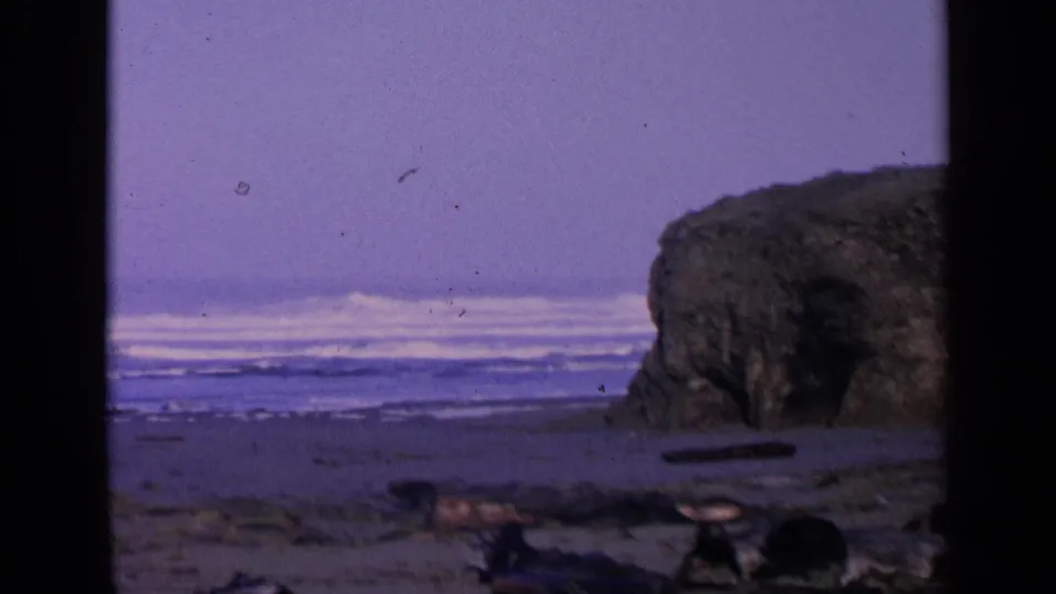 1963 San Francisco-area 8mm frame showing Pacific surf, sandy shoreline, driftwood, and a dark coastal rock formation.