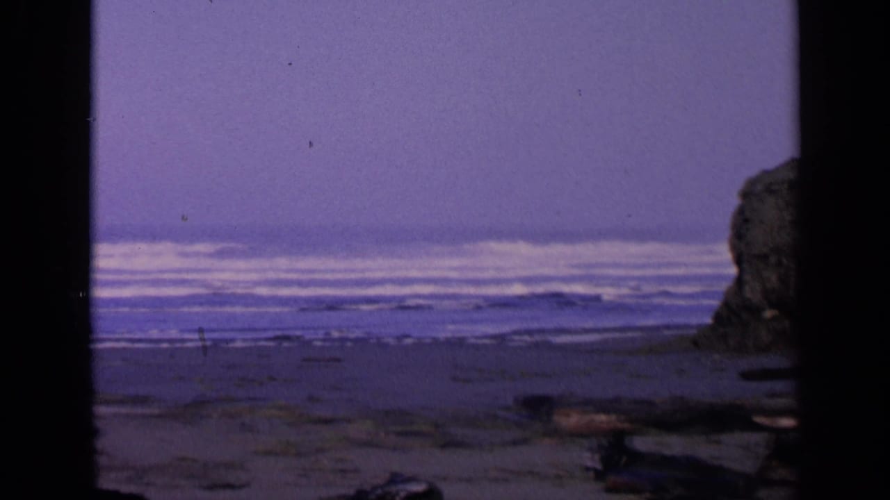 1963 San Francisco-area coastal frame with surf lines, wet sand, driftwood, and a partial rock formation on the right.