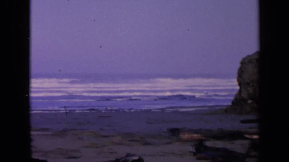 1963 San Francisco-area coastal frame with surf lines, wet sand, driftwood, and a partial rock formation on the right.