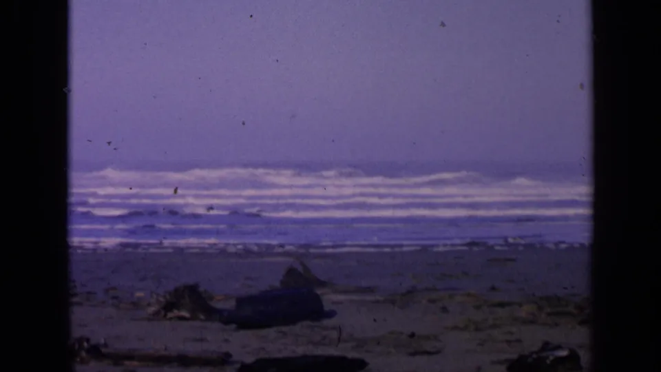 1963 San Francisco-area beach frame with layered Pacific surf, empty sand, driftwood, and visible 8mm film texture.