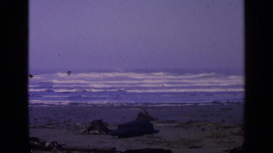 1963 San Francisco coastal frame showing dark sand, driftwood, breaking surf, and a sea stack in the distance.