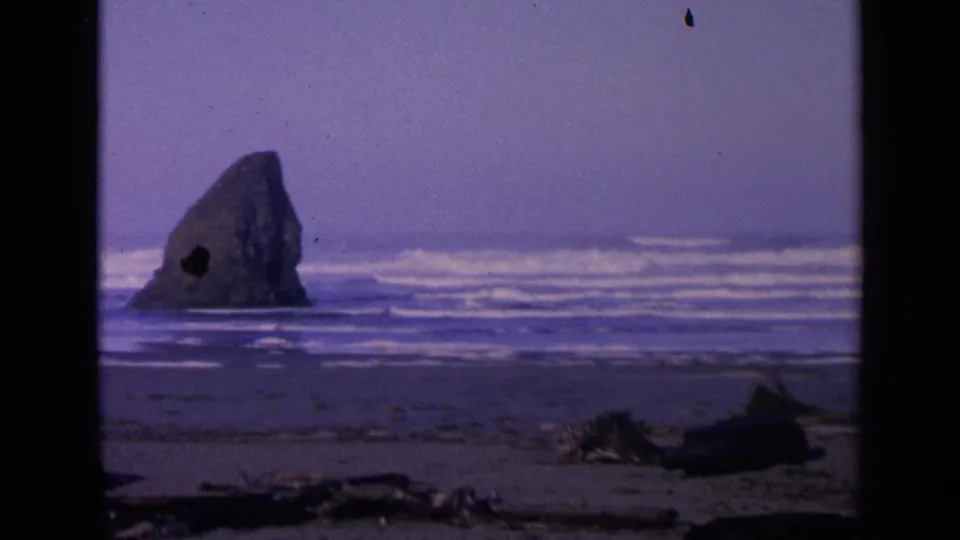1963 San Francisco coastal footage frames a large offshore rock against white surf and dark sand with driftwood below.