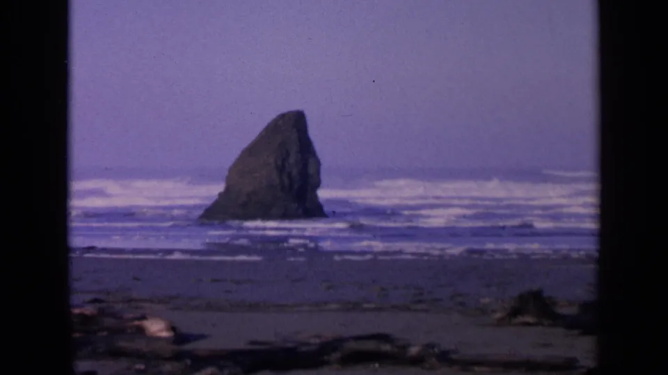 1963 San Francisco shoreline frame centered on a sea stack in rough surf, with dark beach and driftwood in the foreground.