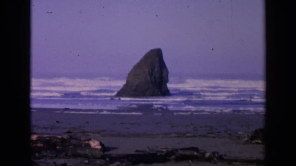 1963 San Francisco-area 8mm frame showing a sea stack beyond wet sand, surf, and driftwood under a purple color cast.