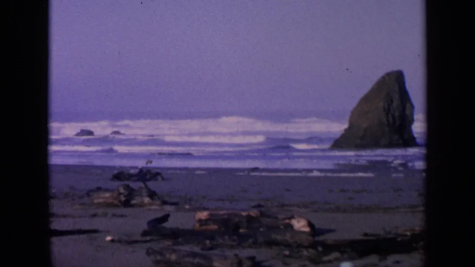 1963 8mm beach frame with the sea stack near the right side, white surf lines, small offshore rocks, and foreground driftwood.