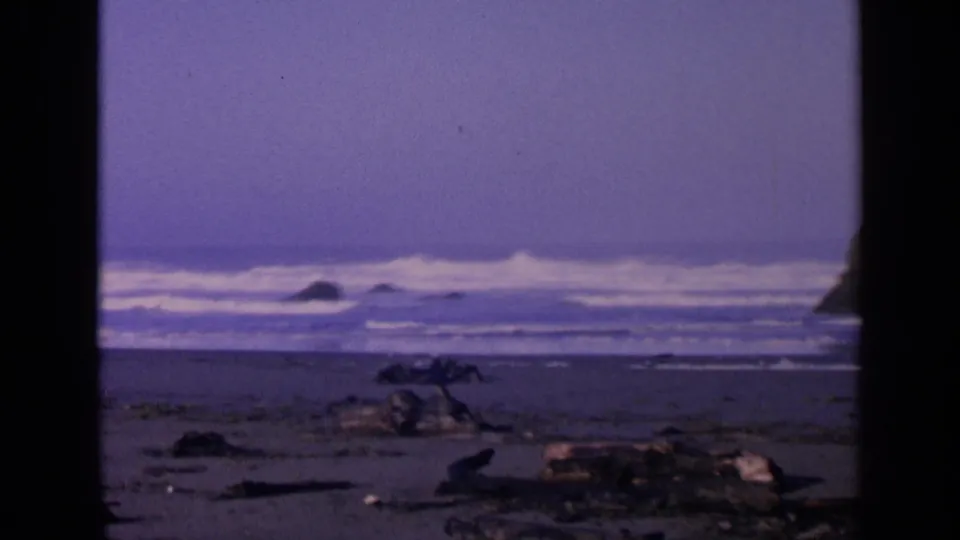 1963 San Francisco 8mm frame showing surf, sand, driftwood, and dark film borders around a quiet beach view.