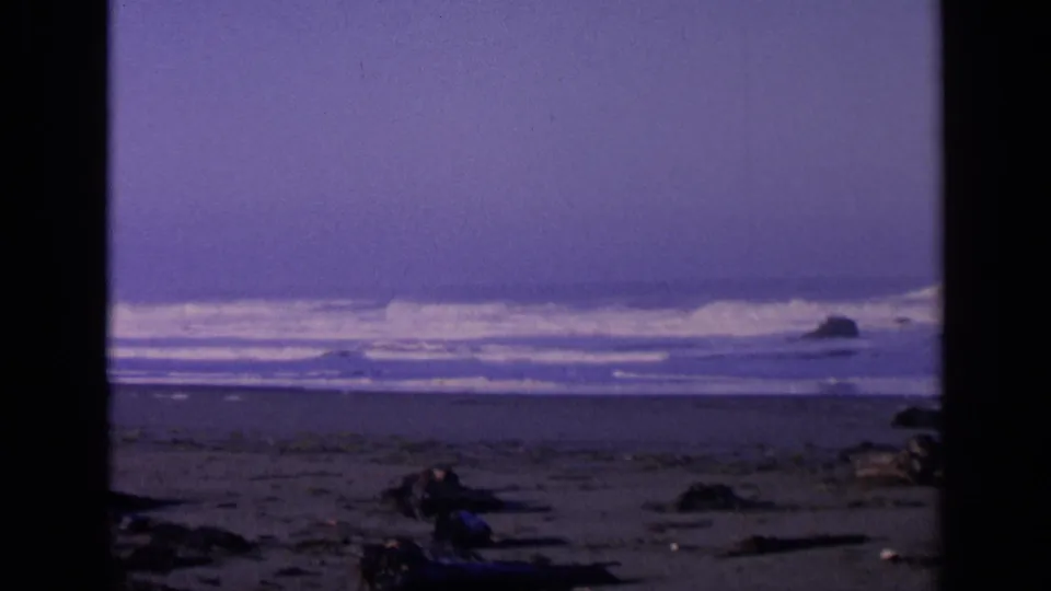 1963 San Francisco home-movie frame showing a wide beach, low surf, offshore rocks, driftwood, and pronounced film borders.