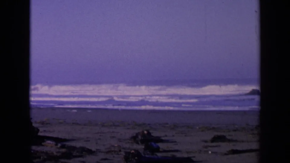 1963 San Francisco beach view on 8mm film, showing low surf, dark driftwood, rocks, and a cool purple-blue sky.