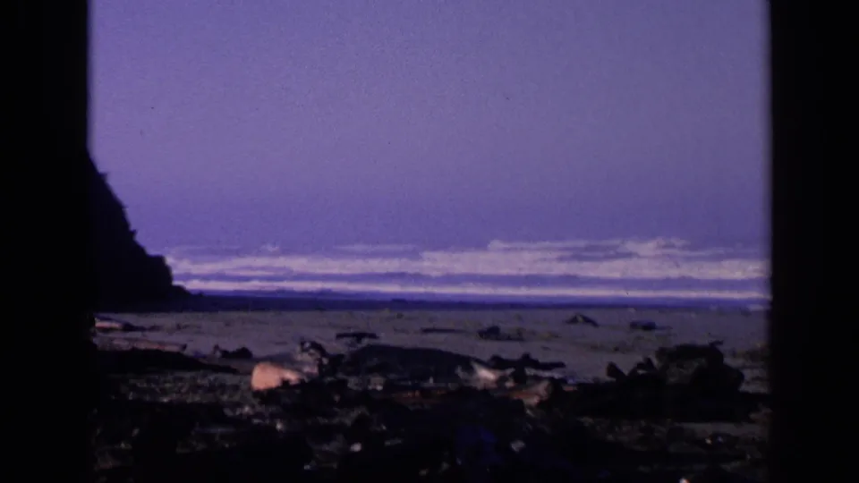 1963 San Francisco-area home-movie frame with a dark cliff edge, rocky foreground, and pale surf offshore.