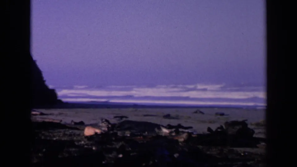 1963 8mm coastal frame with dark rocks, a left-side cliff silhouette, and distant surf under a purple sky.