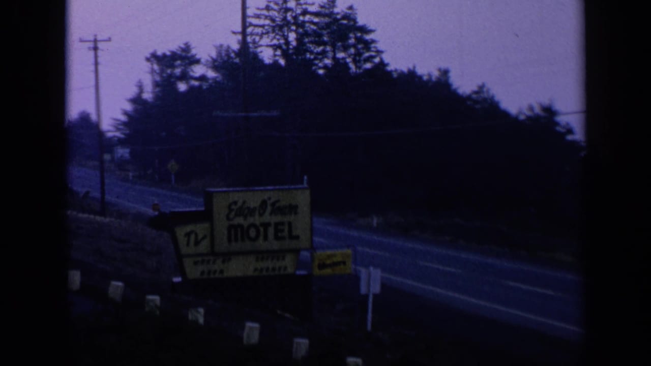 Roadside motel sign beside a two-lane road at dusk with trees and utility poles.