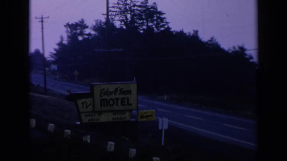 1963 8mm view of an Edge O'Town Motel sign near a quiet roadside, with evergreen trees and a purple dusk sky.