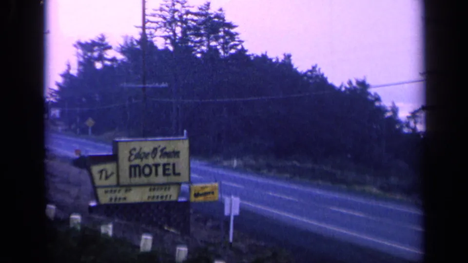 8mm frame showing the Edge O'Town Motel sign, roadside posts, and a quiet road bordered by trees.