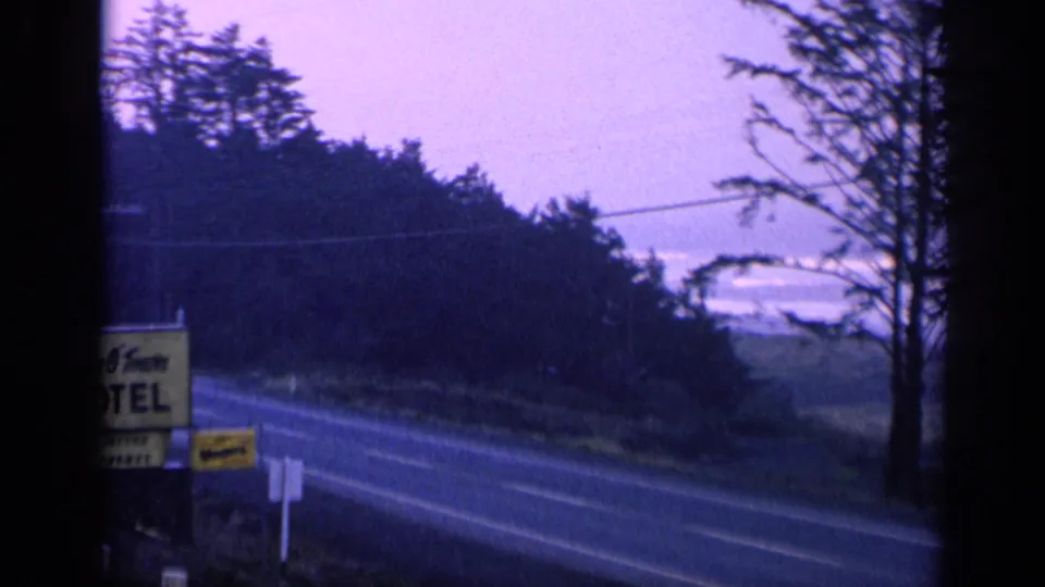 Final 1963 8mm frame with the motel sign partly cut off, a two-lane road, trees, and dusk sky.