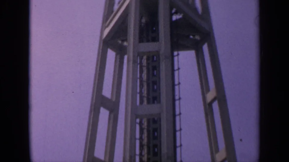Tilted 1963 8mm frame of the Space Needle's lower platform, shaft, and supports against a clear sky.