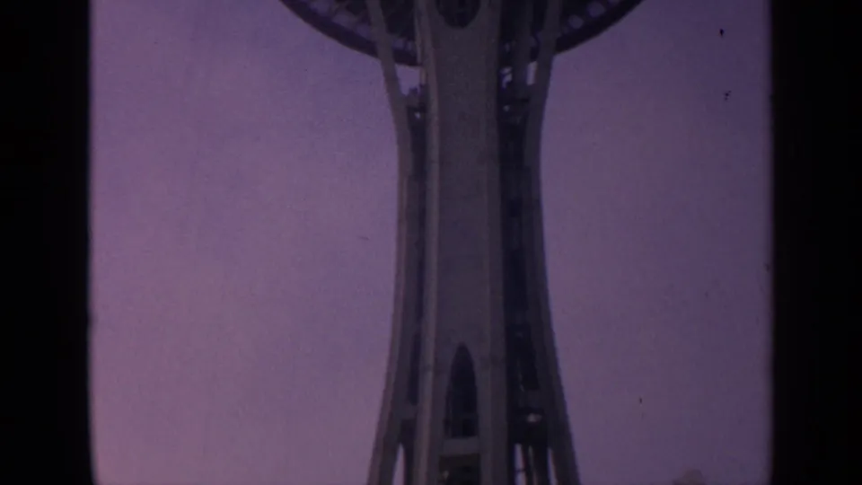 1963 8mm low-angle view of Seattle's Space Needle shaft rising toward the observation deck against a purple sky.
