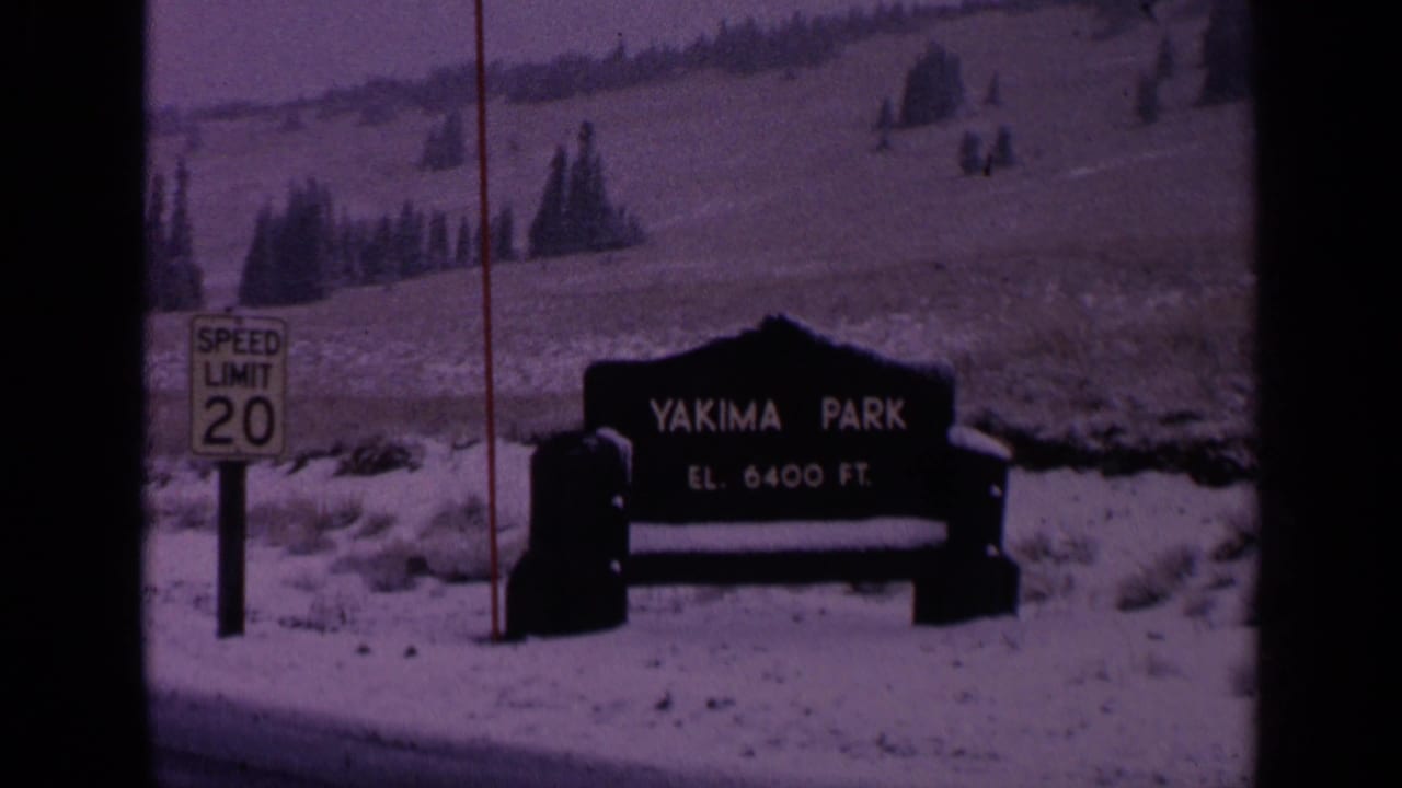 1963 Washington 8mm frame showing the Yakima Park elevation sign in snow, with a speed limit 20 sign and evergreen hillside behind it.