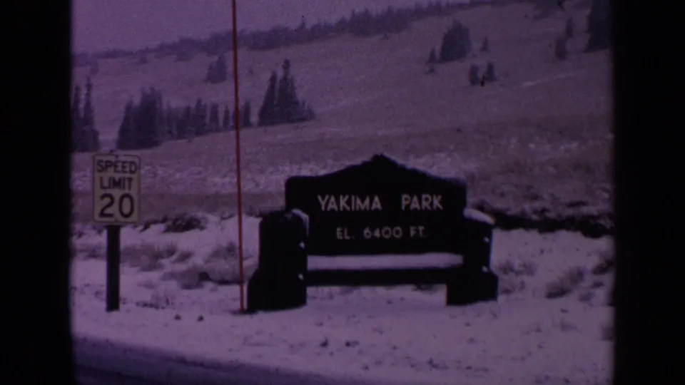 1963 Washington 8mm frame showing the Yakima Park elevation sign in snow, with a speed limit 20 sign and evergreen hillside behind it.