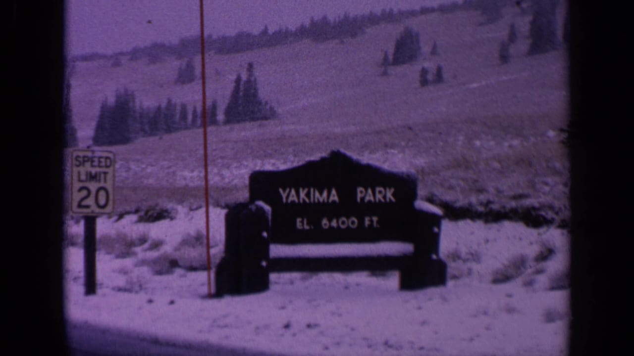 1963 Washington 8mm frame with the Yakima Park sign, elevation marker, road sign, snow-covered ground, and evergreen trees on a hillside.