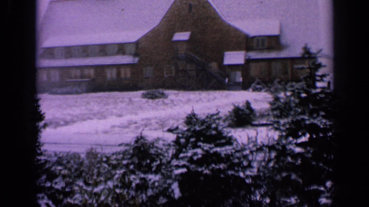 1963 Washington 8mm frame of a winter lodge exterior with snow on the roof, shrubs, evergreens, and visible film-frame edges.