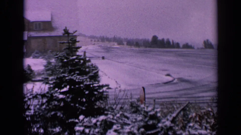 1963 Washington 8mm frame showing a snowy rural field framed by evergreens, foreground shrubs, tire tracks, and a distant building.