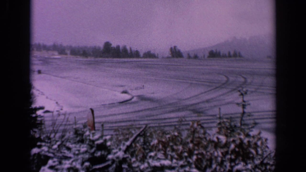 1963 Washington 8mm view across an open snow field with foreground brush, vehicle tracks, distant trees, and faint hills beyond.