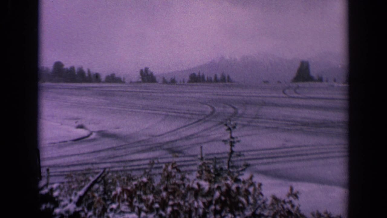 1963 Washington home-movie frame showing a wide snow-covered field, layered tire tracks, foreground shrubs, and faint mountains in the distance.