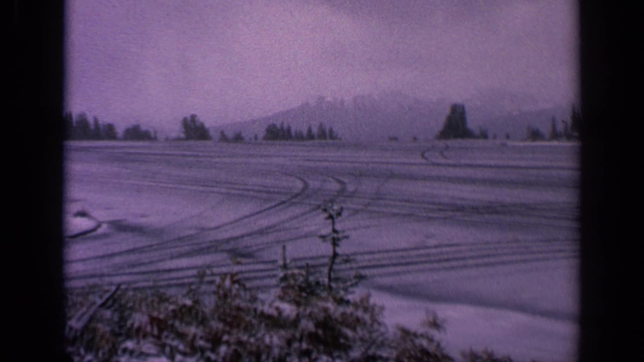 1963 Washington 8mm frame showing a snowy rural field, tire tracks, distant mountains, evergreens, and foreground shrubs under an overcast sky.
