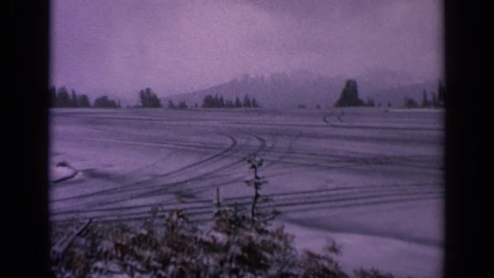 1963 Washington 8mm frame showing a snowy rural field, tire tracks, distant mountains, evergreens, and foreground shrubs under an overcast sky.