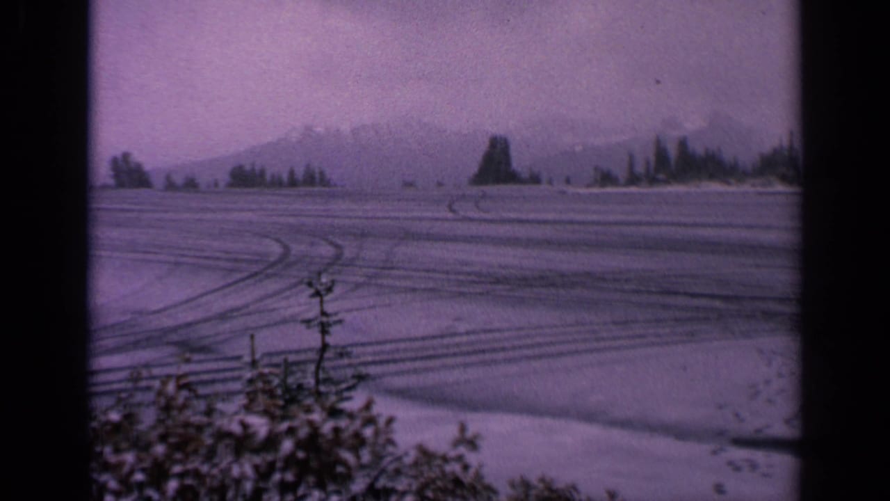 Open snowy field with tire tracks, low mountains, tree line, and dark vertical film edge.