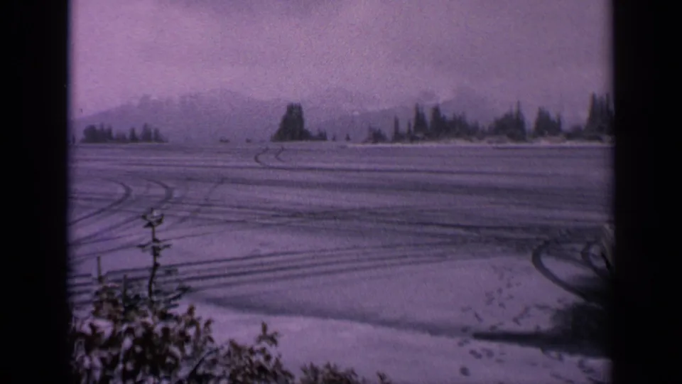 1963 Washington 8mm frame showing tire tracks across a snow-covered field, distant evergreens, mountain silhouettes, foreground brush, and muted winter light.
