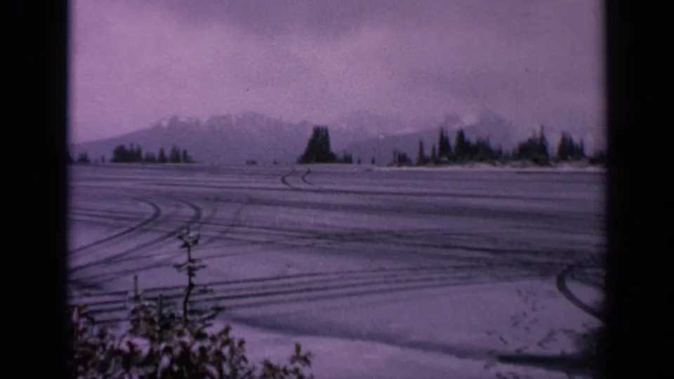 1963 Washington 8mm frame of an empty winter field with curved tire tracks, snow-laden foreground shrubs, distant mountains, and scattered evergreens.