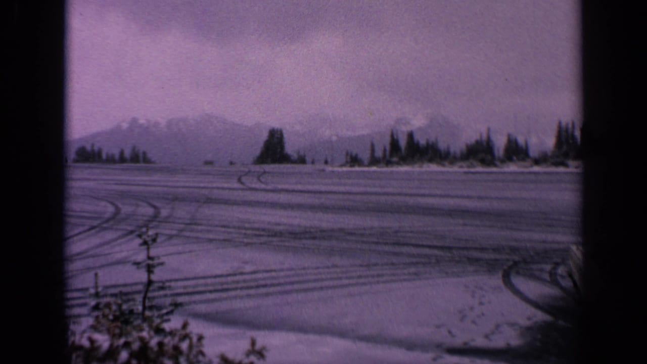 Snow-covered open lot with tire tracks, shoreline trees, distant mountains, and part of a white coupe at right.