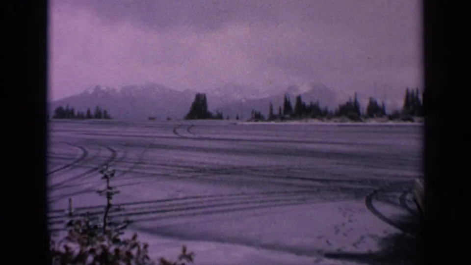 1963 Washington 8mm frame showing a snowy lot with tire tracks, dark shoreline trees, distant mountains, and the edge of a white coupe.