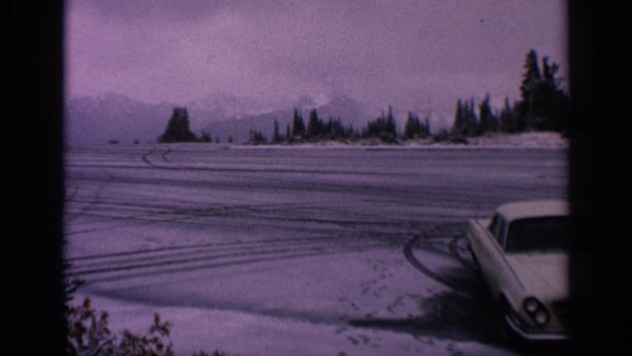 White coupe parked at the edge of a snowy lot with tire marks, trees, mountains, and dark film borders.
