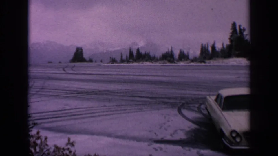 1963 Washington home-movie frame of a white coupe parked near a snowy open lot with curved tire tracks and distant tree-lined mountains.