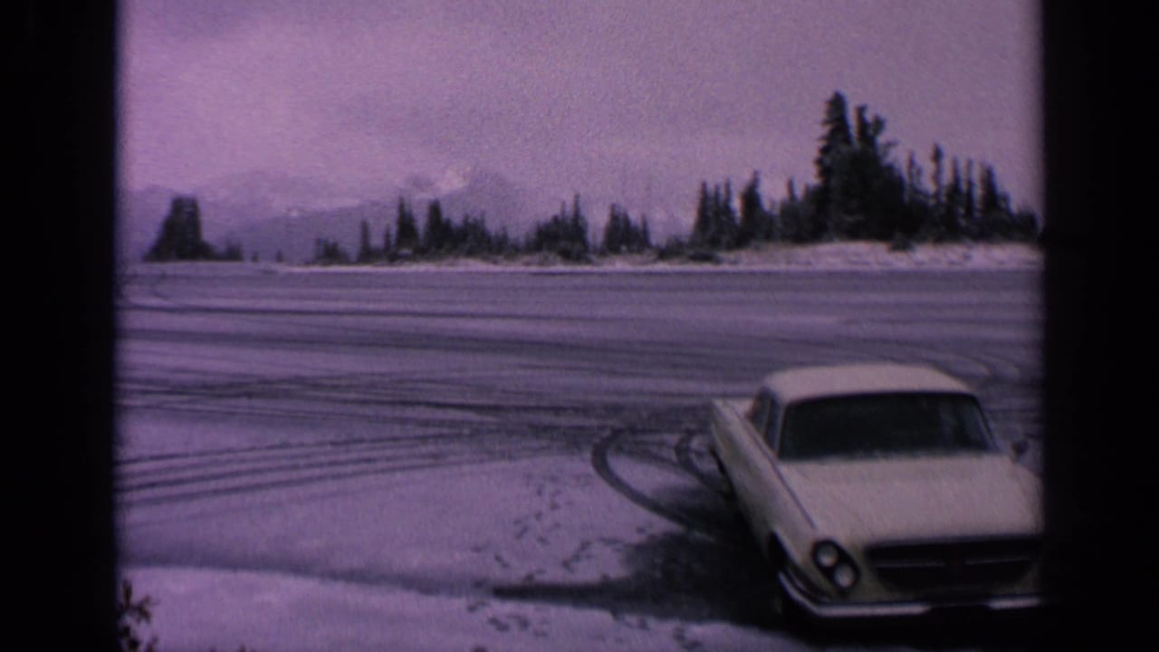 1963 Washington 8mm frame showing the front of a white coupe beside sweeping tire tracks on snow, with trees and mountains in the distance.