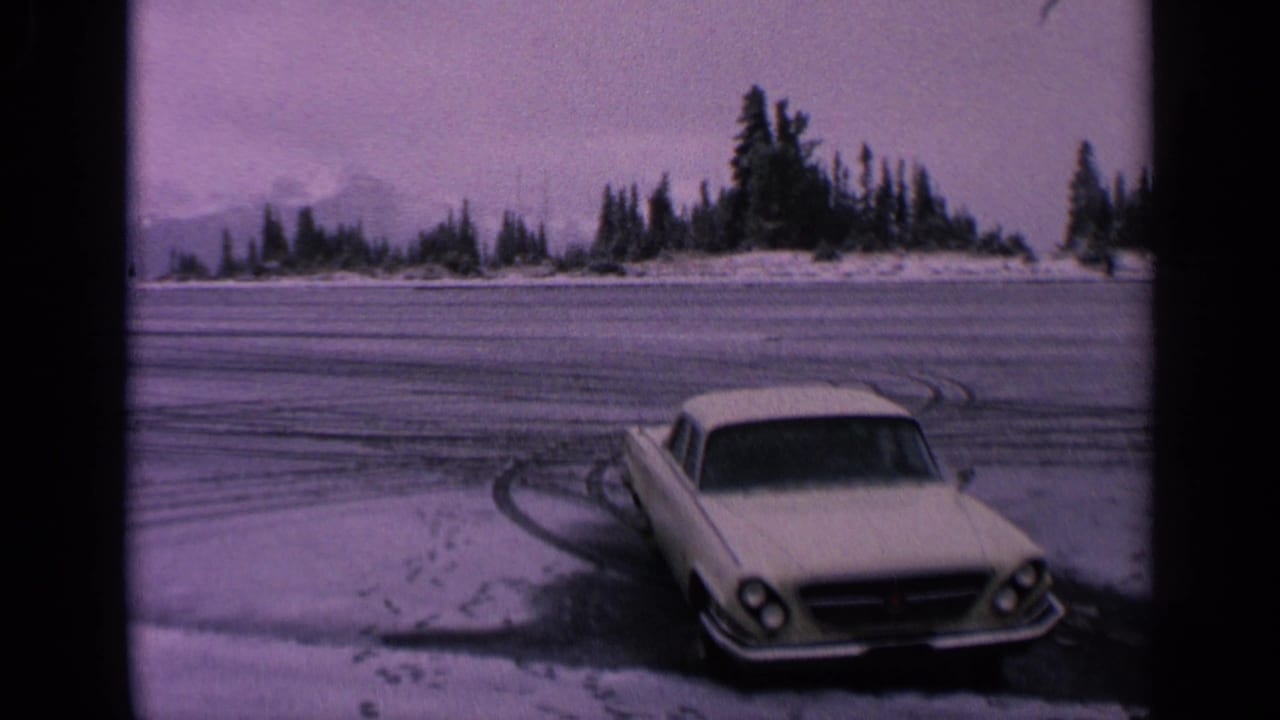 1963 Washington home-movie frame of a white coupe parked beside a snow-covered lot, with tire tracks leading toward trees and distant mountains.
