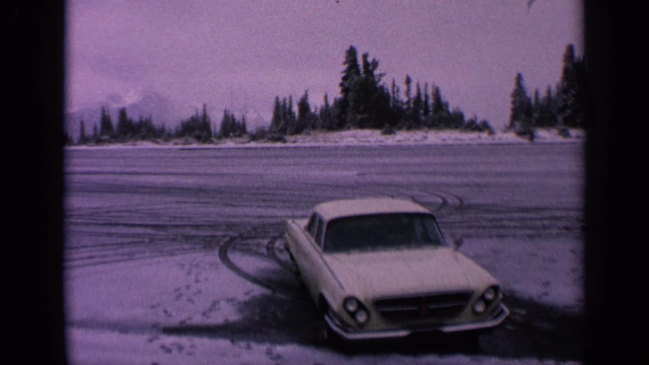 1963 Washington home-movie frame showing a white car on a frozen roadside with tire tracks, snow patches, and evergreen trees beyond.